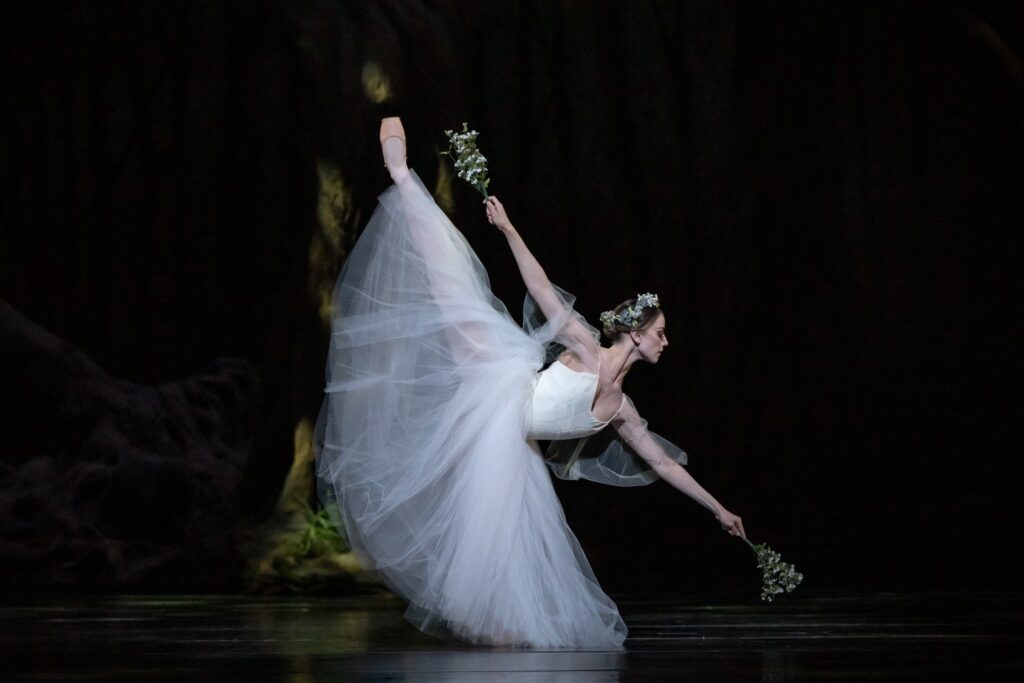 Tyler Dontelli, wearing a long white tutu and a wrather of white flowers in her hair, does a first arabesque penché on a darkened stage. She holds a small bouquet of white flowers in both hands.