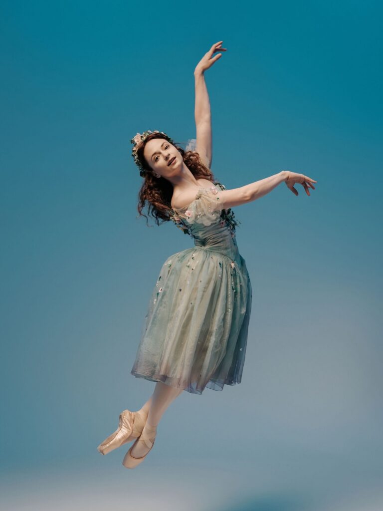 Chantelle Pianetta jumps up, her feet in pointe shoes and crossed at the ankle, and lifts her arms to third arabesque as she arches slightly with her upper body. She wears a long blue tutu with a filmy overlay and puffed, off-the-shoulder sleeves. Her red hair is worn down, crowned with a tiara, and she jumps in front of a blue backdrop.