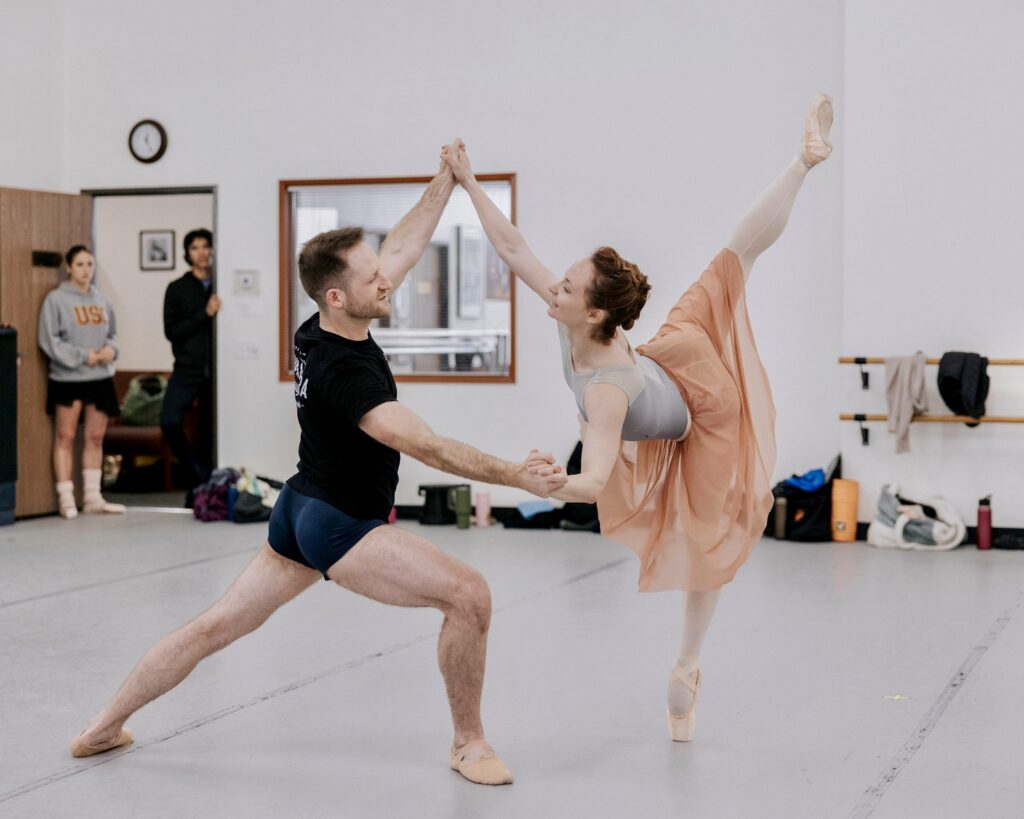 Chantelle Pianetta and Preston Swovelin are shown rehearsing in a dance studio. Swovelin is in a deep lunge facing Pianetta, who is in arabesque penché en pointe. Their hands are clasped, forming a diamond in the center of the frame and they smile at each other. Pianetta wears a light grey leotard with a flowy salmon-pink skirt. Swovelin wears navy shorts and a black T-shirt.