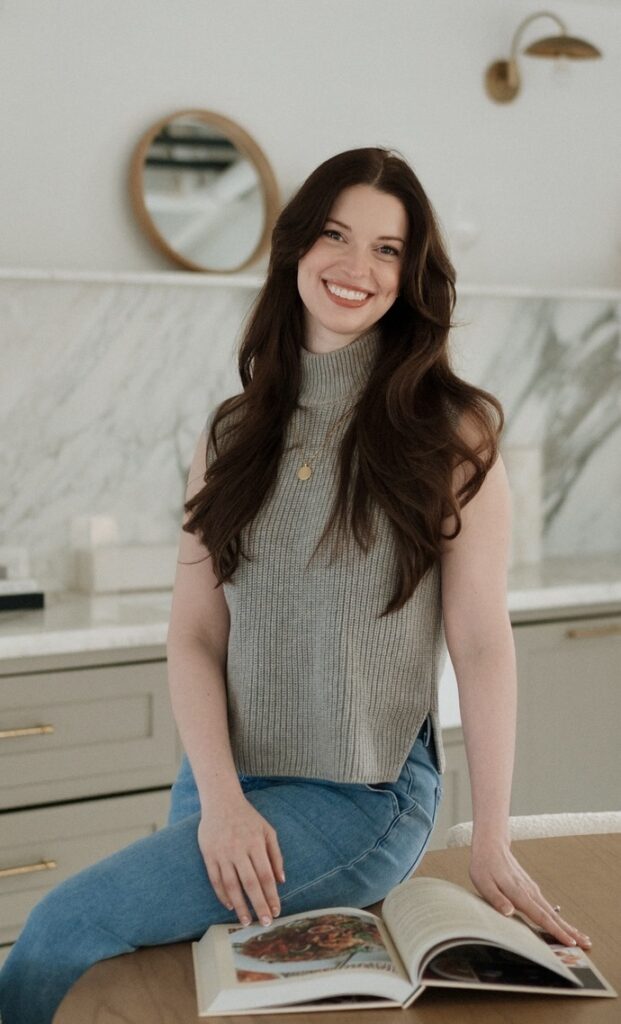 Kelsey Fyffe is shown sitting sideways on the edge of a table and twisting her upper body towards the camera. She wears blue jeans and a sleeveless taupe turtleneck, and her long dark hair down. She smiles at the camera as she touches a large open book on the table.