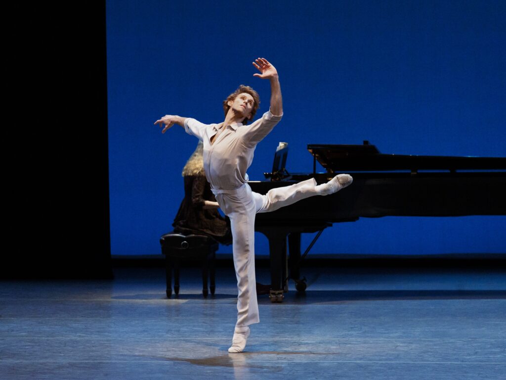 Adrian Danchig-Waring does a piqué attitude derrière in croisé onstage, lifting his right leg behind. He holds his right arm out to the side and lifts his left arm up, his palm facing out as he looks over his left shoulder. He wears a white shirt and pants, and a woman plays on the piano in the background.