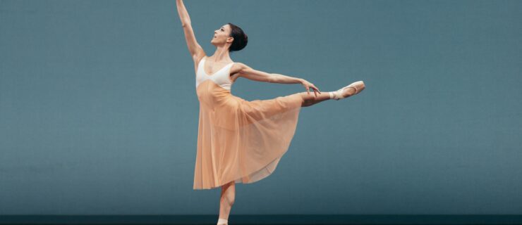 Katherine Barkman does a piqué first arabesque towards stage right. Her costume is a long pink and white dress and pointe shoes, and she performs in front of a blue backdrop.