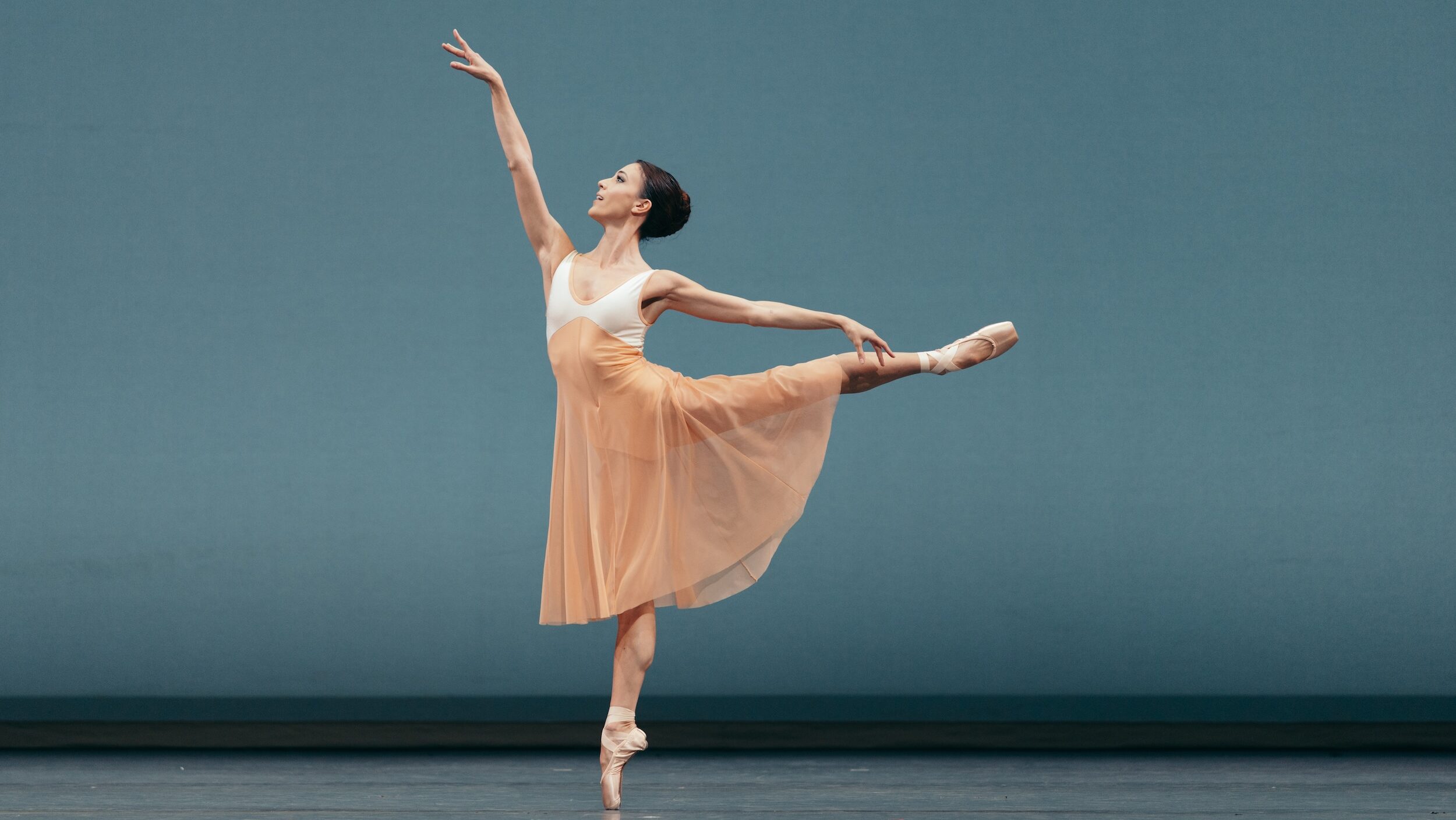 Katherine Barkman does a piqué first arabesque towards stage right. Her costume is a long pink and white dress and pointe shoes, and she performs in front of a blue backdrop.