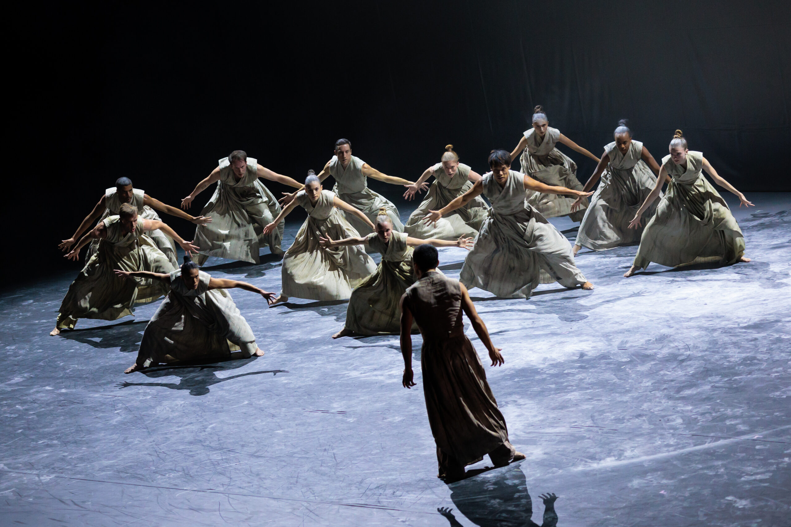 A group of dancers wearing neutral-colored costumes with large, baggy skirts faces another dancer whose back is toward the audience. The group pliés in second position, bending their upper bodies forward slightly and spreading their arms wide. their expressions are intense, and the lone dancer stands before them in a strong stance.