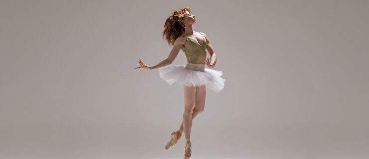 Chantelle Pianetta is shown mid-air against a light background. She wears skin-tone pointe shoes and has one leg in coupé behind the other. Pianetta wears a light green leotard and white tutu. She has a calm expression, her face in profile and hair loose. Pianetta holds her arms low, one rounded in front of her, above the tutu, and the other behind.