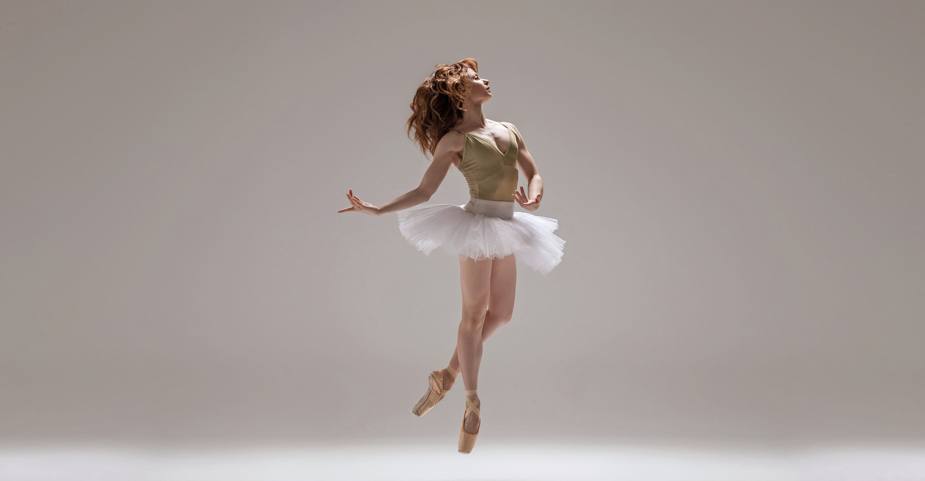 Chantelle Pianetta is shown mid-air against a light background. She wears skin-tone pointe shoes and has one leg in coupé behind the other. Pianetta wears a light green leotard and white tutu. She has a calm expression, her face in profile and hair loose. Pianetta holds her arms low, one rounded in front of her, above the tutu, and the other behind.