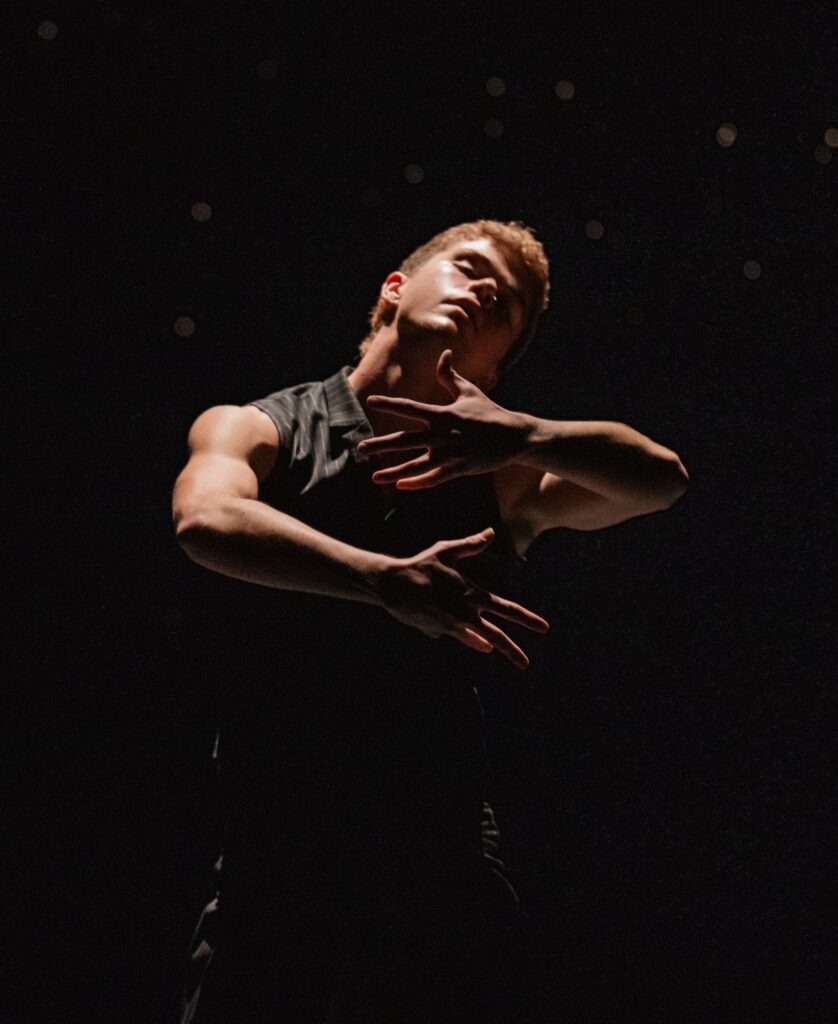 Clay Houston is shown from the knees up, spotlighted from above on a darkened stage. He wears a drak sleeveless top and holds his arms in front of him as if in an embrace as he closes his eyes, tilting his head to his left.