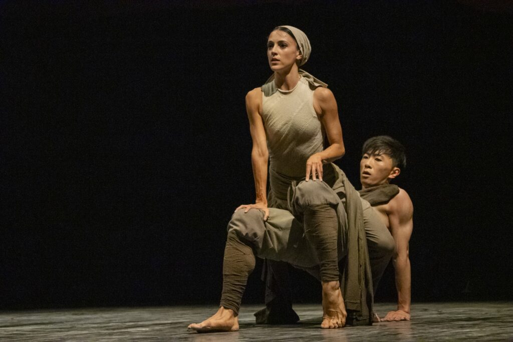 Katherine Barkman and Wei Wang dance a pas de deux onstage. Wei sits back and pushes up on his hands and feet as Brakman crals on top of him, arching her back up. They wear gray costumes, and Barkman has a gray scarf tied around her head.