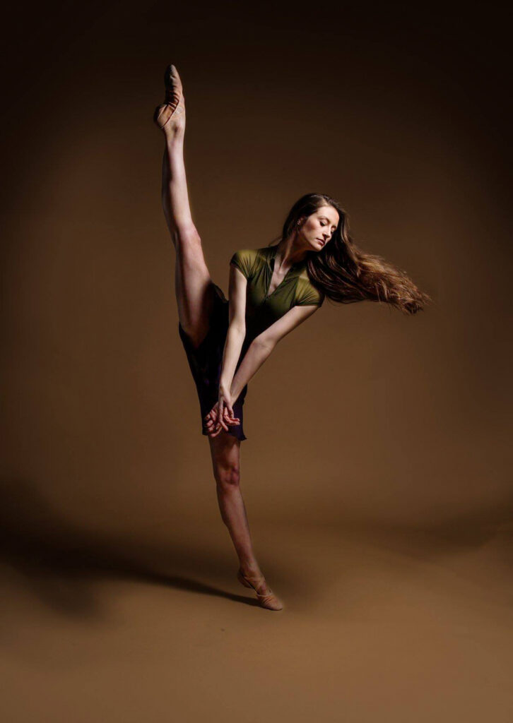 Deanna Stanton clasps her hands low with straight arms as she tilts and battements her right leg straight up. She wears a short-sleeved olive-colored leotard and a dark ballet skirt.