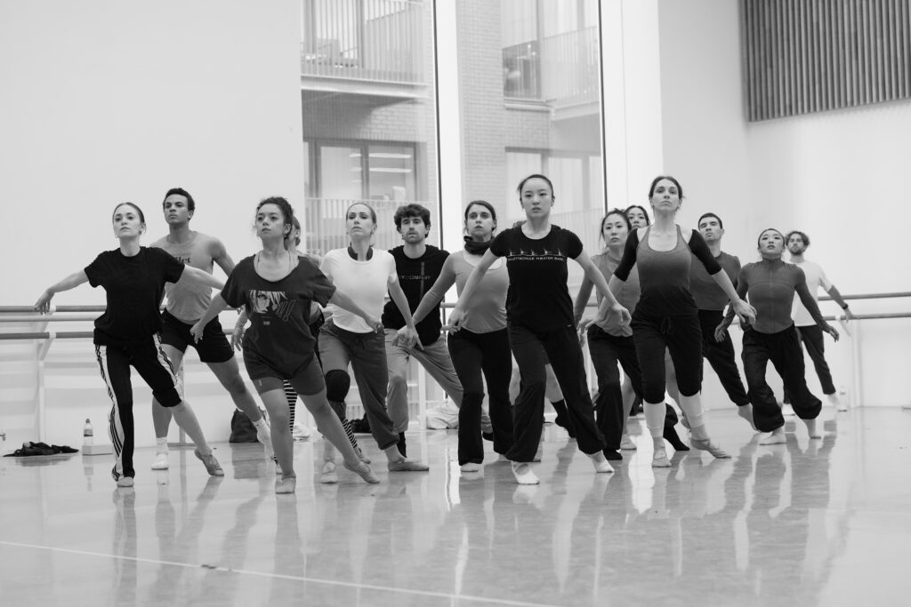 A black-and-white photo of a large group of dancers arranged in a clump in the corner of a ballet studio. They lunge forward with their back legs extended, their back feet on demi pointe, as they open their chests wide and hold their arms down and behind them.