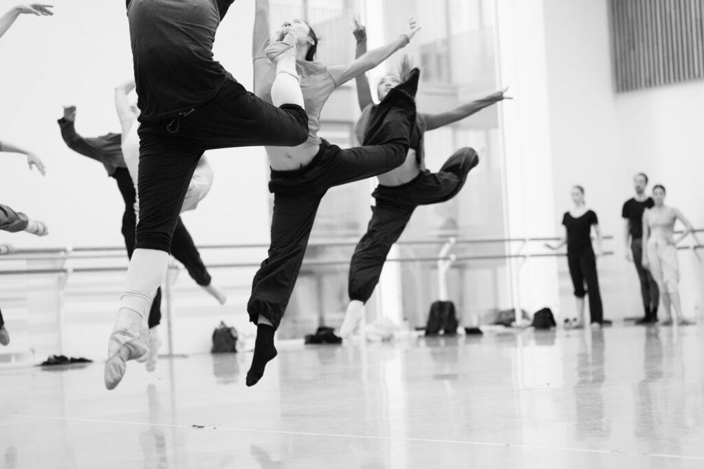 A black-and-white photo of dancers rehearsing in a large ballet studio. On the left, dancers in two rows sauté forward. with their back legs in attitude derriere, their arms flung upward in a high arabesque position.