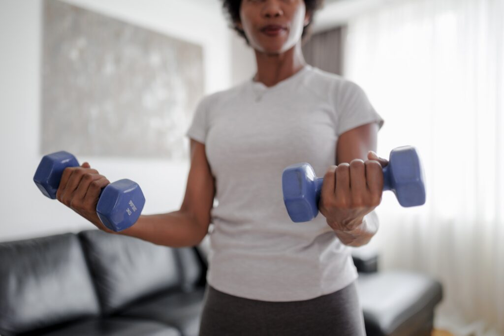 A woman is shown, from her hips to the bridge of her nose, exercising in a brightly lit living room. She is wearing a light gray short-sleeved shirt and dark grey bottoms and lifts a purple dumbbell in with each hand.