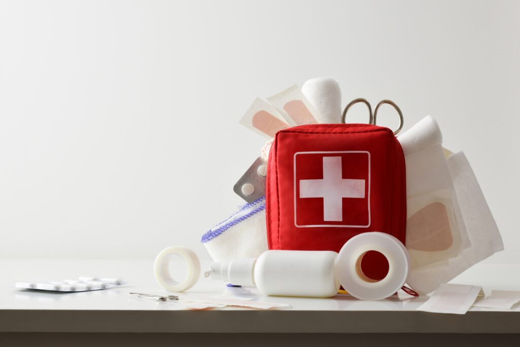 A red travel first-aid bag with a large white cross on the front is propped up on a white table, full of bandages, rolls of tape, scissors, and other first aid supplies. Some of the supplies spill out onto the table.