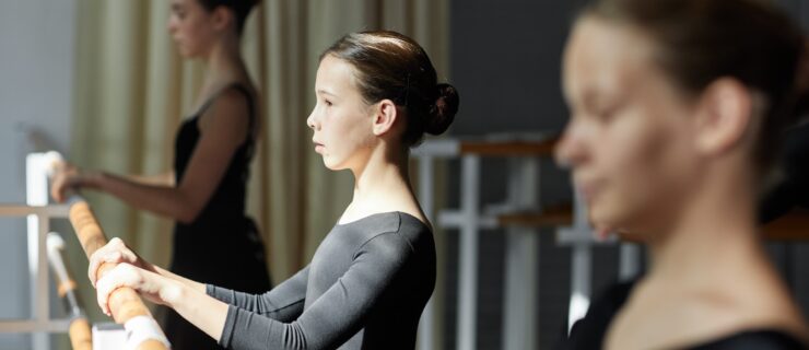 Side view of concentrated teenage girl in a long-sleeved black leotard facing the barre during ballet class in sunlit studio. She stands between two older teenage dancers.