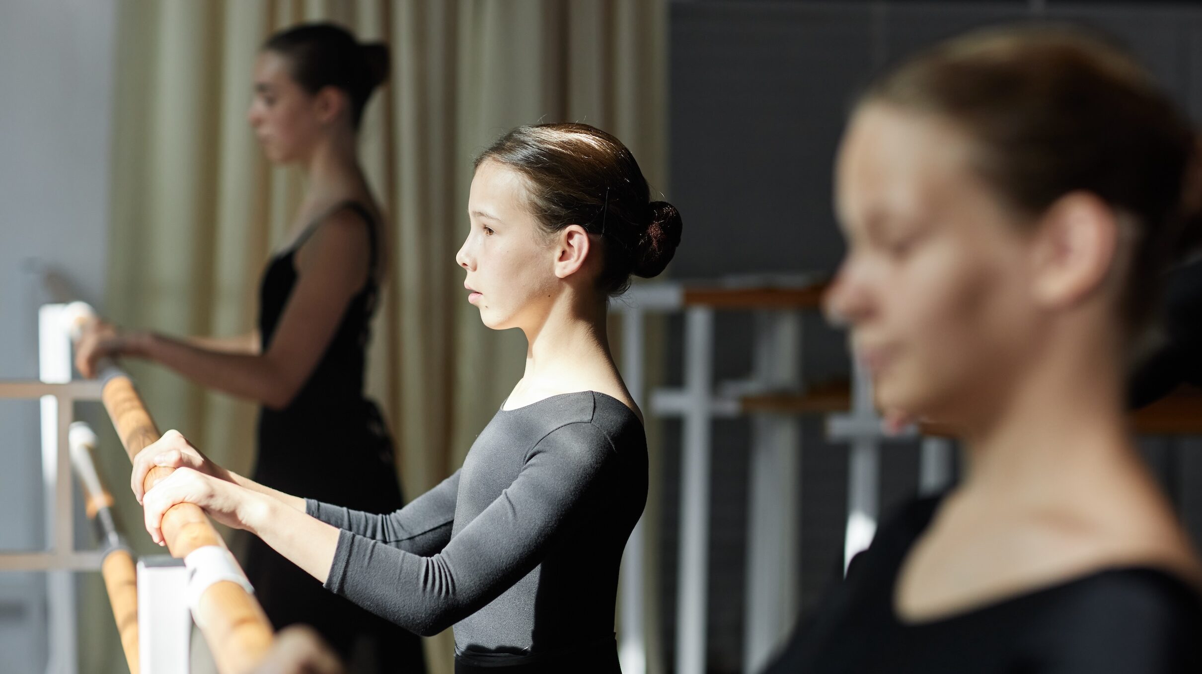Side view of concentrated teenage girl in a long-sleeved black leotard facing the barre during ballet class in sunlit studio. She stands between two older teenage dancers.