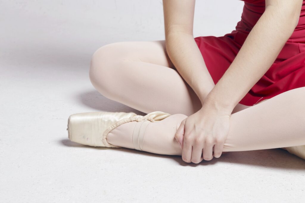Close up of a ballerina from the ribcage down, sitting with her legs crossed on a white floor. She wears a red leotard and skirt with pink tights and has her pointe shoe half off, freeing her heel. She holds her ankle.