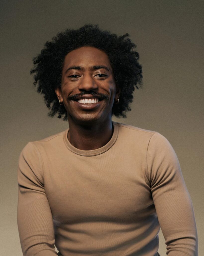 A headshot of Houston Thomas from the waist up. He wears a beige long-sleeved shirt and hoop earrings. The background is a muted green. Thomas smiles into the camera.