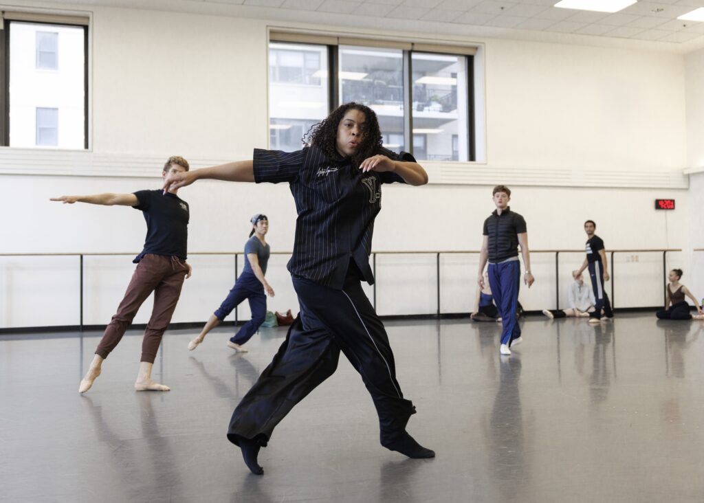 Jennifer Archibald moves swiftly in a ballet studio, crossing her right leg over her left as she swings her arms to the right. Three men in practice clothes stand behind her and watch, trying to mimic her. Archibald wears dark pants and a matching three-quarter-sleeve shirt.