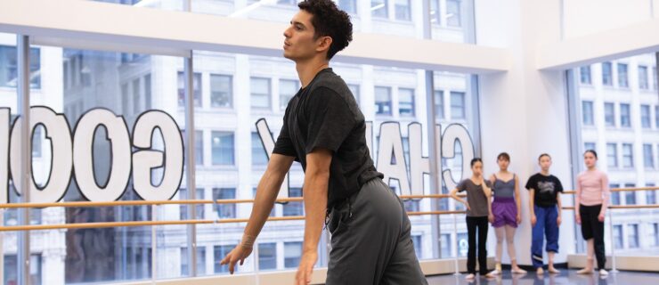 In a large dance studio with huge windows, Xavier Núñez flows through movement during a rehearsal. He wears a dark t-shirt and gray workout pants. Four young female dancers stand in the corner of the room, watching him as he demonstrates.