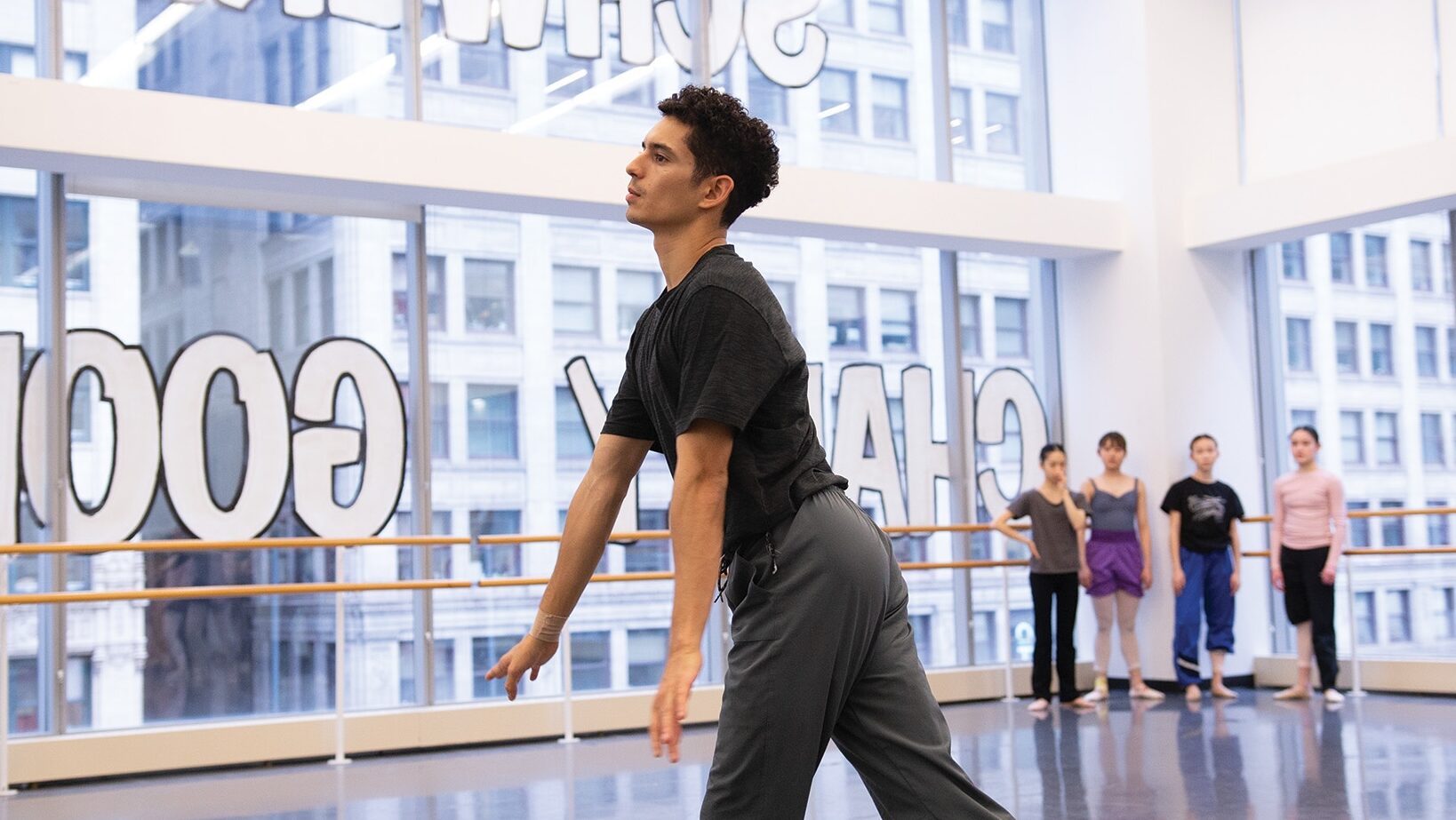 In a large dance studio with huge windows, Xavier Núñez flows through movement during a rehearsal. He wears a dark t-shirt and gray workout pants. Four young female dancers stand in the corner of the room, watching him as he demonstrates.