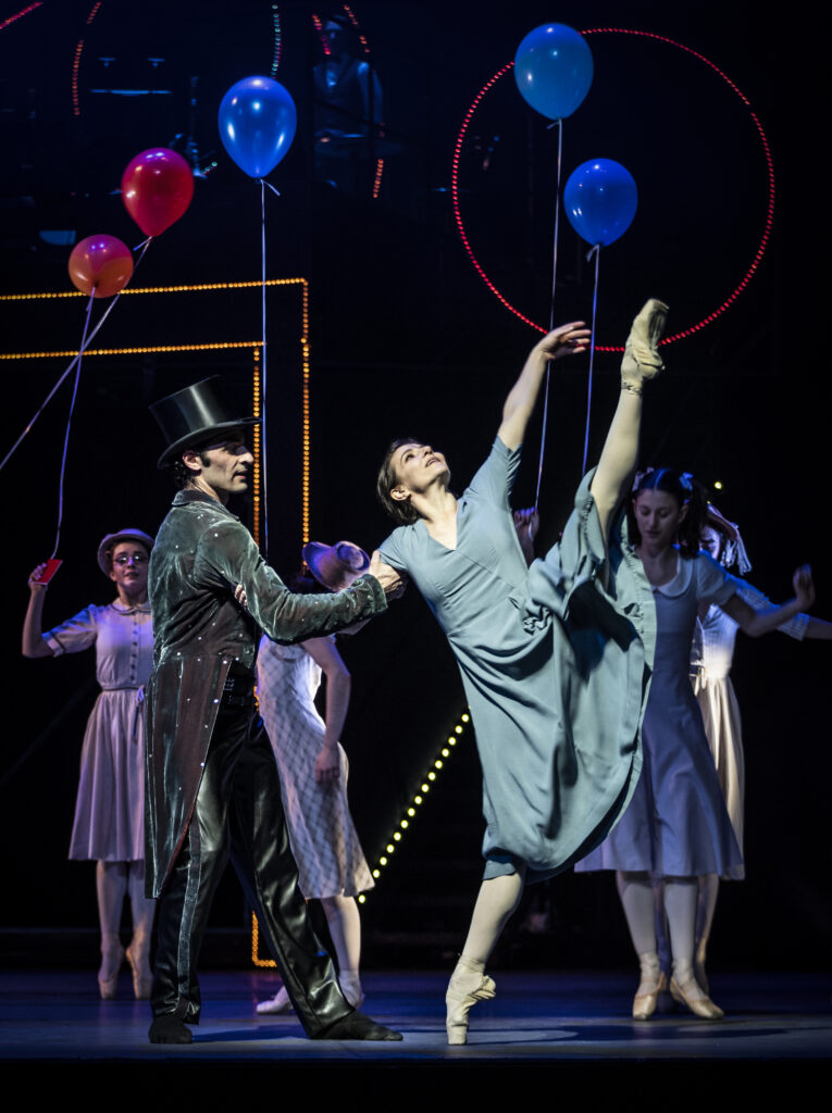 On a dark stage with colorful lights and balloons, a carnival caller in a velvet coat and a top hat clasps arms with a dancer in a light blue dress who battements one leg high on pointe. She lifts her other arm up to follow the lifted leg's line, her eyes turned up toward the ceiling. The caller looks at her, his back to the audience, as carnival-goers dance in the background. 