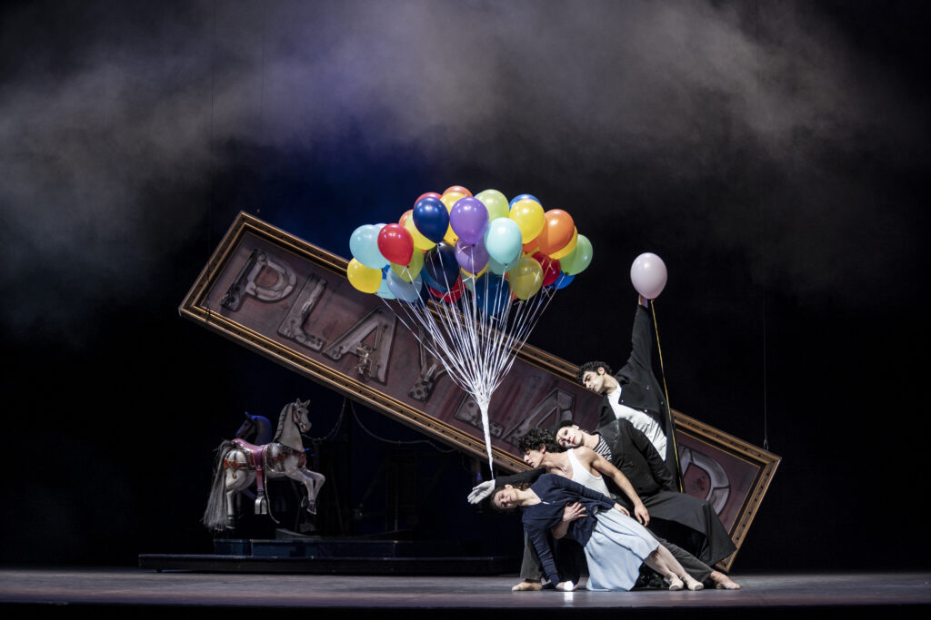 Onstage in front of a dark backdrop with fog, carousel horses, and a "Playground" marquee, four dancers stand in a slight diagonal line. Each person holds onto the one in front of them with one arm as they lean to the left in a "wave" effect. The dancer in the back holds one balloon high; a mime-like character in front of him reaches out one arm, to which a large bundle of balloons is tied. In front, the protagonist holds his love interest around her waist. 