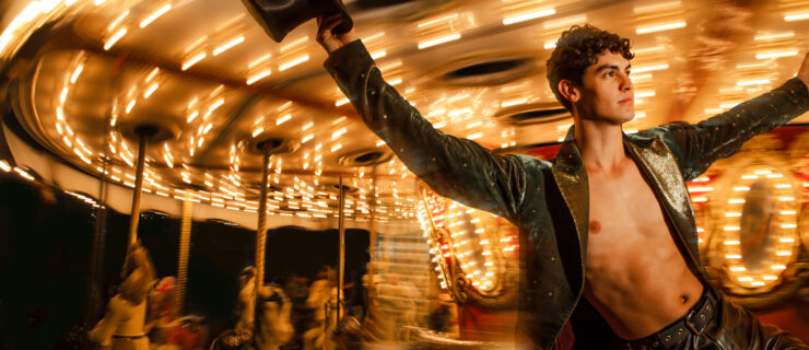 A dancer in leather pants and an open leather jacket holds a top hat while hanging onto a carousel pole. The carousel moves with a blurred effect as the dancer looks off into the distance calmly.
