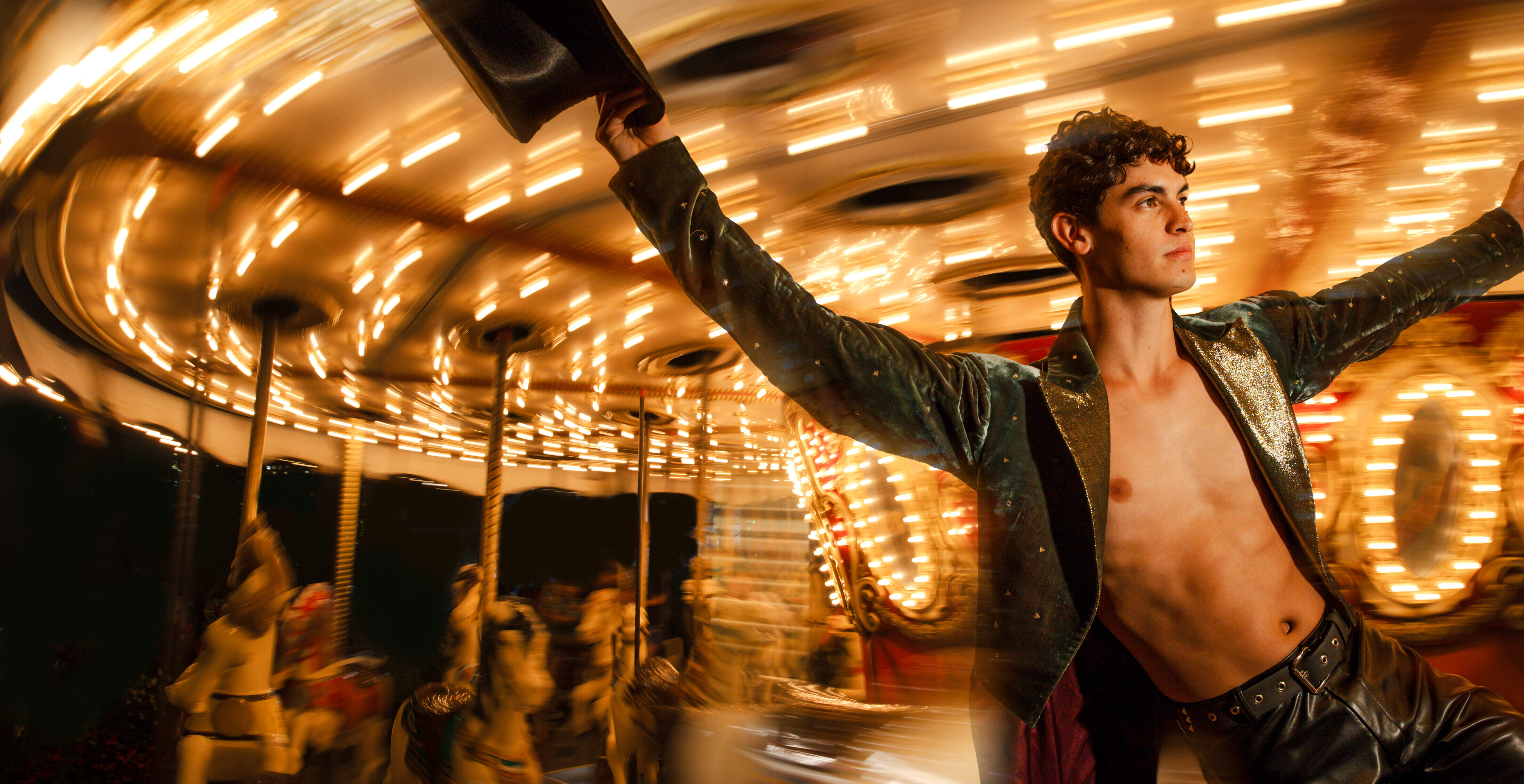 A dancer in leather pants and an open leather jacket holds a top hat while hanging onto a carousel pole. The carousel moves with a blurred effect as the dancer looks off into the distance calmly.