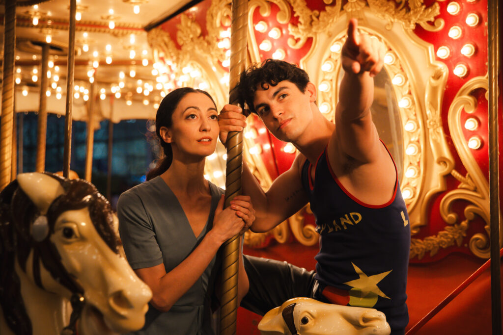 Two dancers ride a carousel with gold lights and white horses. They both hold onto a gold pole belonging to one of the horses. The dancer on the right, one leg propped up, points off camera, and his partner follows his point with her eyes.