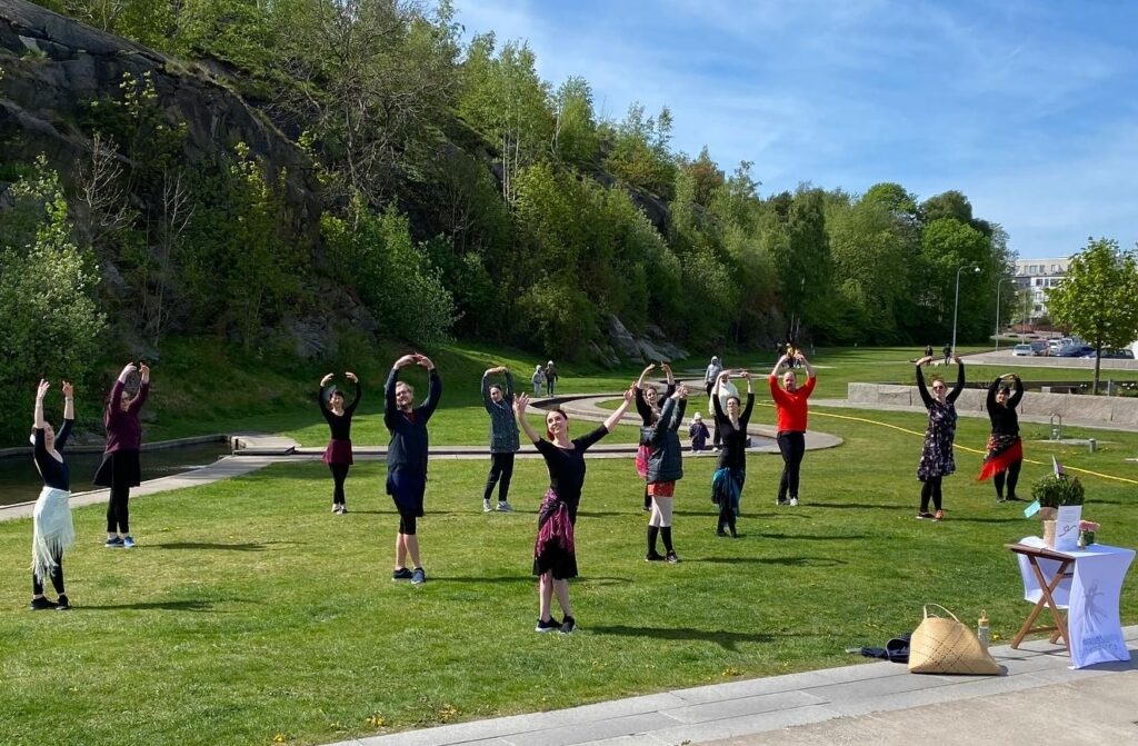 Lori Boggan is shown leading her class of dancers in a grassy park with a forested rock formation behind them. Standing in front of the group, she wears black clothing with a pink wrap skirt and black sneakers. The dancers behind her are similarly dressed in loose clothing and sneakers. Boggan and all the dancers are standing in croisé fifth position with their arms in high fifth and their heads turned over their shoulder. Boggan is opening her arms with a smile.
