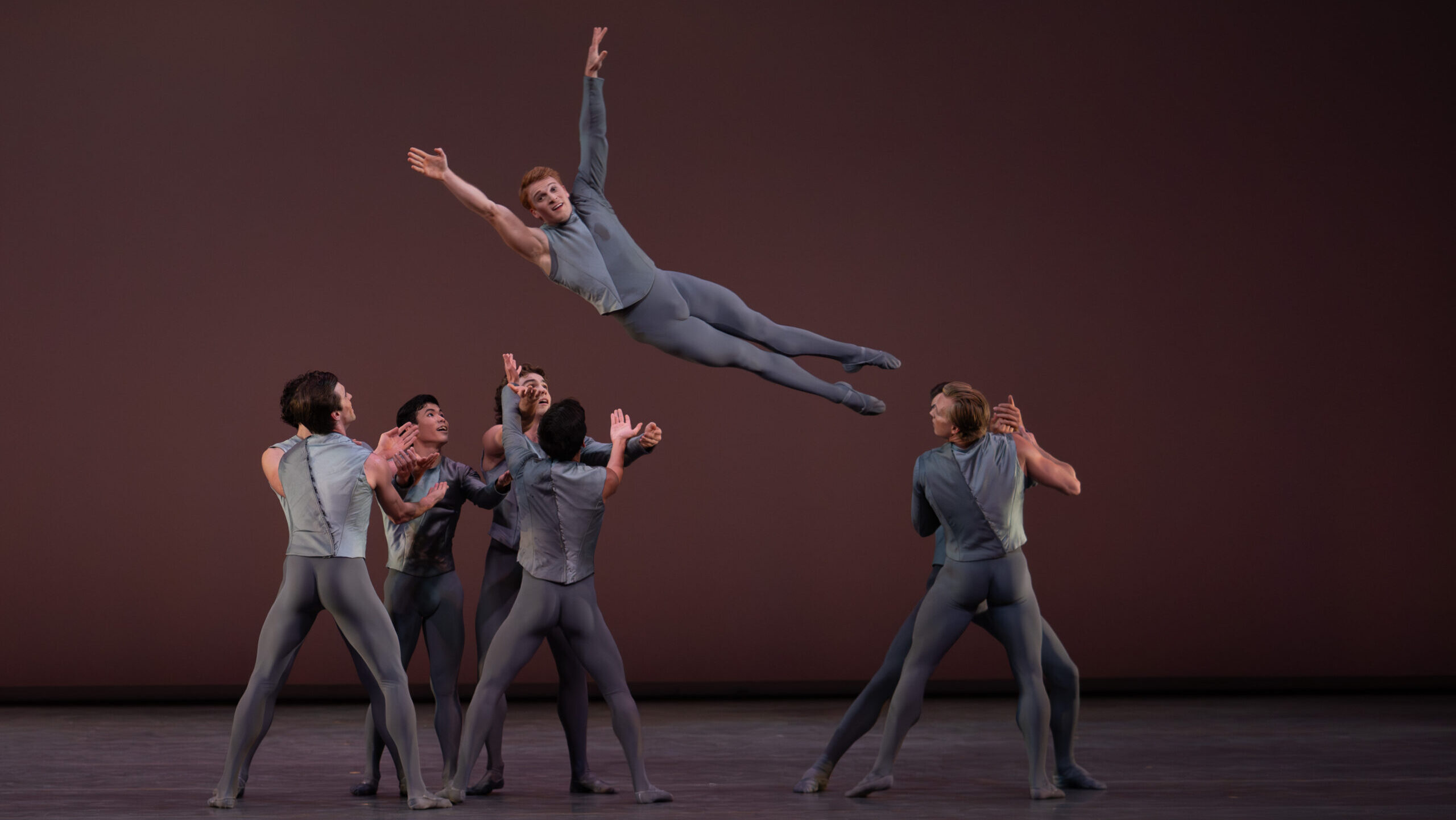 A group of eight male dancers are shown mid-lift onstage. One dancer is midair above the heads of the others, facing the audience with his legs straight and arms in a V above is head, while the rest of the dancers prepare to catch him. All wear medium grey tights and a grey two-toned top that is long-sleeved on one arm and sleeveless on the other.