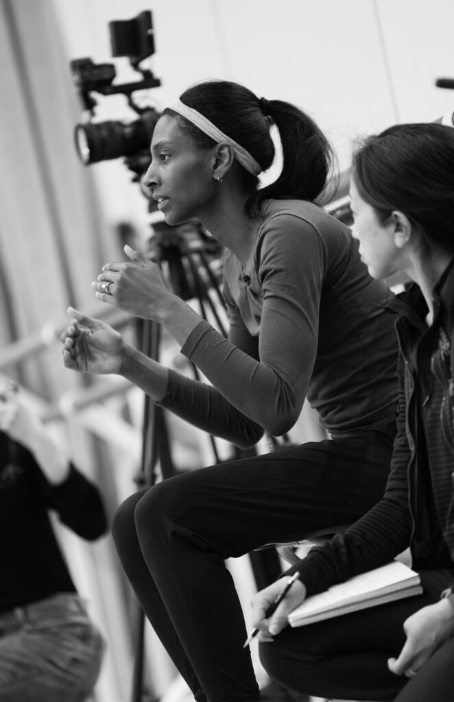 A black-and-white photo of Micaela Taylor leading rehearsal in a ballet studio. She sits on a stool and lifts her arms gently, elbows bent, while speaking to dancers who are out of frame. A rehearsal assistant sits next to her with a pad and pencil.