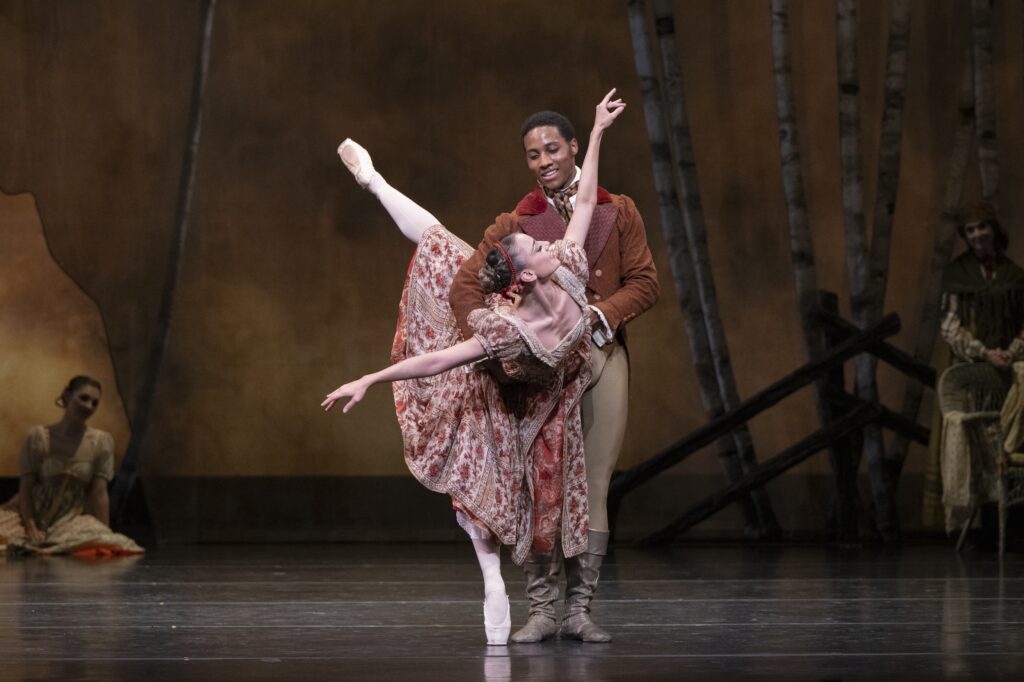 Tyler Donatelli and Eric Best perform a pas de deux onstage. Donatelli, costumed in a long red and gold brocaded dress, does a penché in croise, her arms out. She looks back towards Best, who wears an orange russet jacket and tan tights and boots. He holds her by the waist and looks down at her adoringly with a smile.