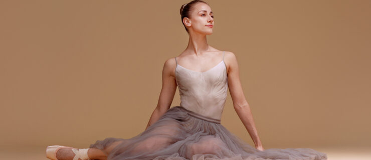 Tyler Donatelli sits on the floor in front od a beige backdrop. Her hair is tightly pulled back, and she wears a long, gray tutu, a light pink camisole leotard with a faint gray pattern, and pointe shoes. She tucks her left leg underneath her and extends her right leg out a bit, and looks out towards her left with a faraway gaze and slight smile.