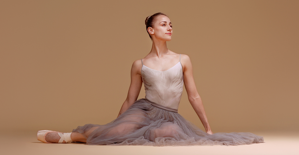 Tyler Donatelli sits on the floor in front od a beige backdrop. Her hair is tightly pulled back, and she wears a long, gray tutu, a light pink camisole leotard with a faint gray pattern, and pointe shoes. She tucks her left leg underneath her and extends her right leg out a bit, and looks out towards her left with a faraway gaze and slight smile.