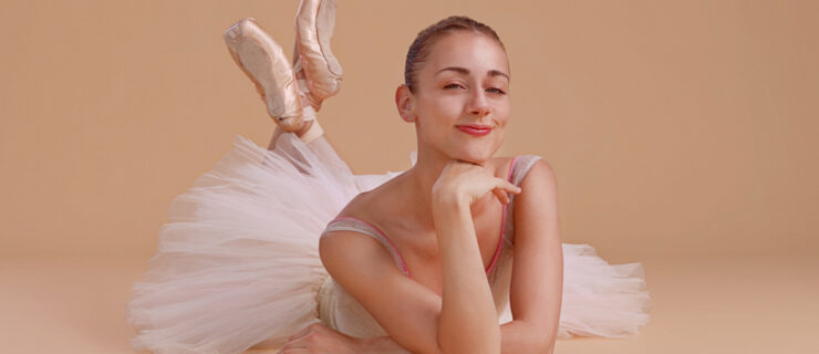 Tyler Donatelli lays down on her stomach in front of a beige photo backdrop. Wearing a white tutu, she rests on her elbows and props her chin up with the back of her right hand. Her feet are lifted behind her, crossed at the ankles, and she smiles cheekily at the camera.