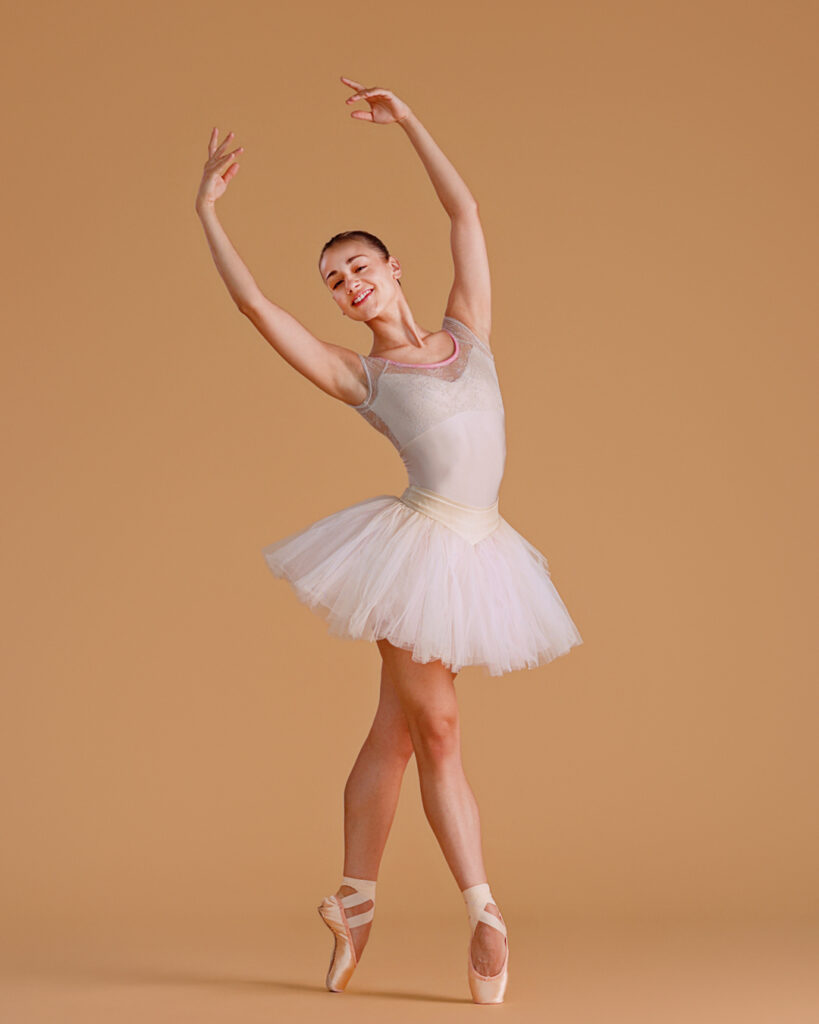 Tyler Donatelli poses in fourth position croisé on pointe with her arms in high fifth. She wears her hair pulled back in a bun and a white tutu, and poses in front of a beige backdrop.