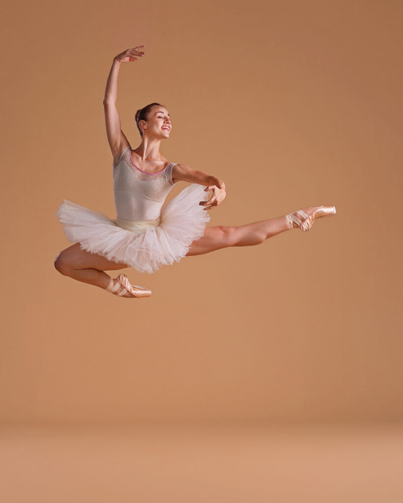 Tyler Donatelli performs an Italian pas de chat with her arms in high third in front of a beige backdrop. She wears a white tutu and pointe shoes, and her hair is pulled back in a bun. She looks out over her left arm with a joyous smile.