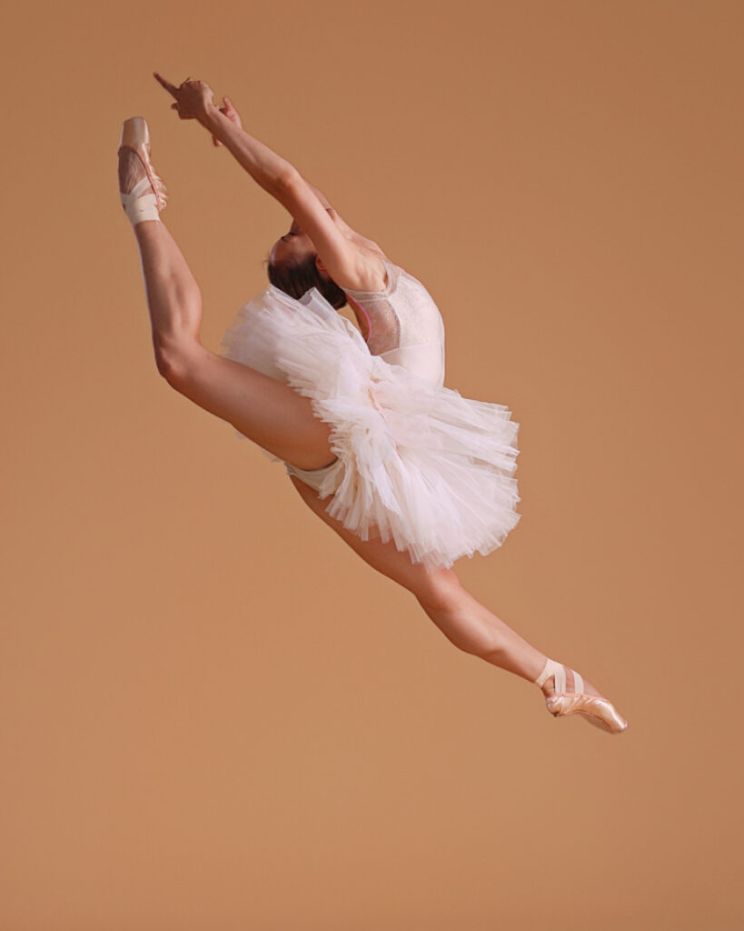 Tyler Donatelli jumps up into a large sissone, her legs split and her back leg in attitude, almost kicking her head, which is thrown back. She wears a white tutu and pointe shoes, and jumps in front of a beige backdrop.