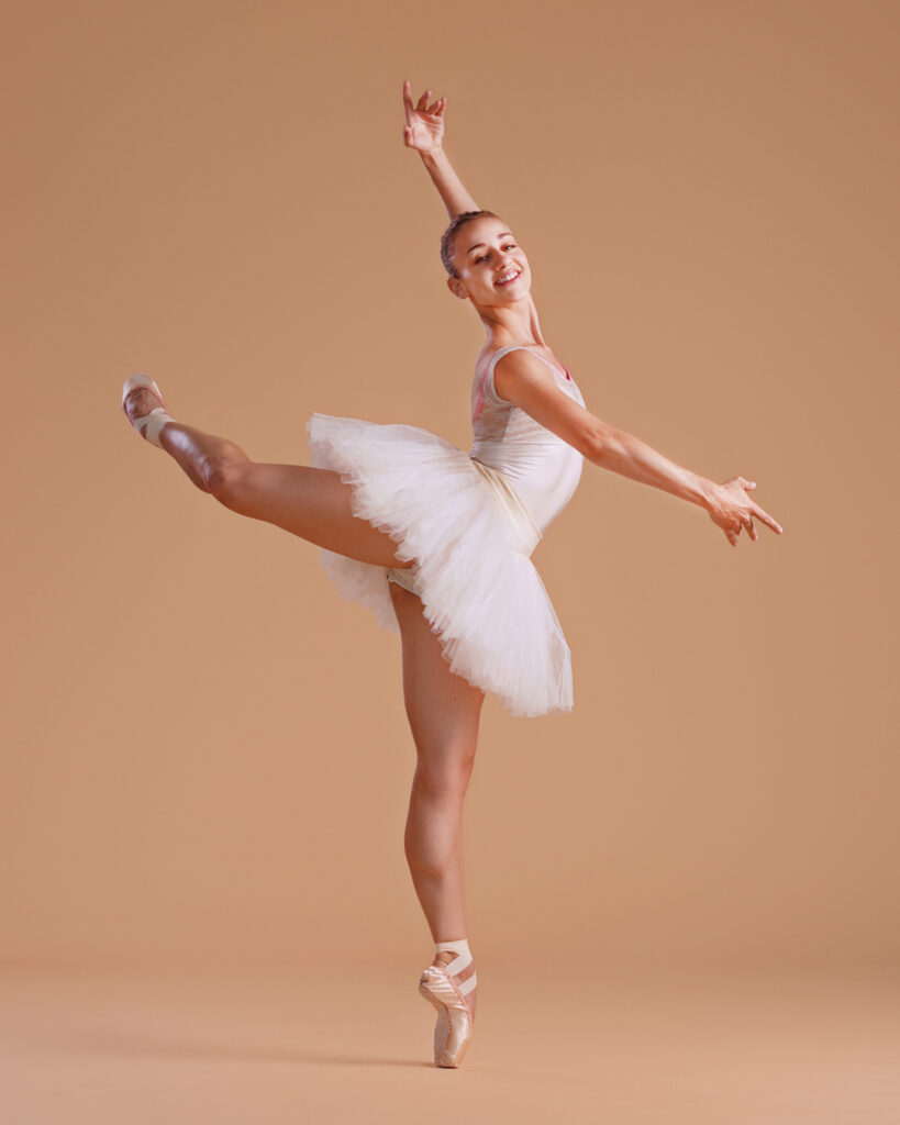Tyler Donaltelli wears a white tutu and pointe shoes and poses with her right leg raised in attitude derriere effacé. She lifts her left amr up and her right arm is curved in front of her, and she looks out towards the camera with a large smile.