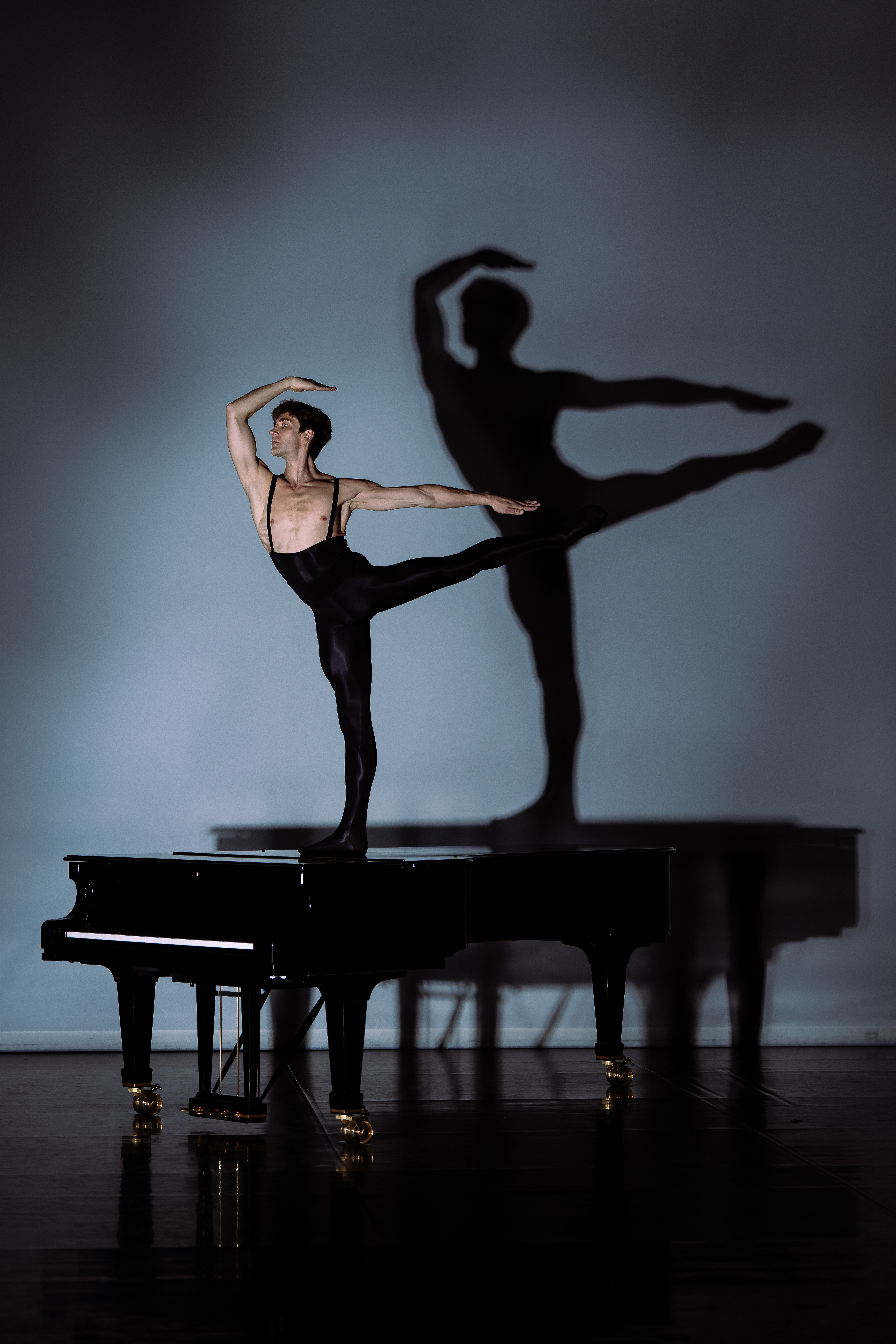 Friedmann Vogel is shown in onstage, holding arabesque on top of a grand piano. Vogel wears black tights and frames his face with one arm in an angular fifth position, his other arm extended straight behind him. His shadow creates a prominent silhouette of his pose against the light grey backdrop.