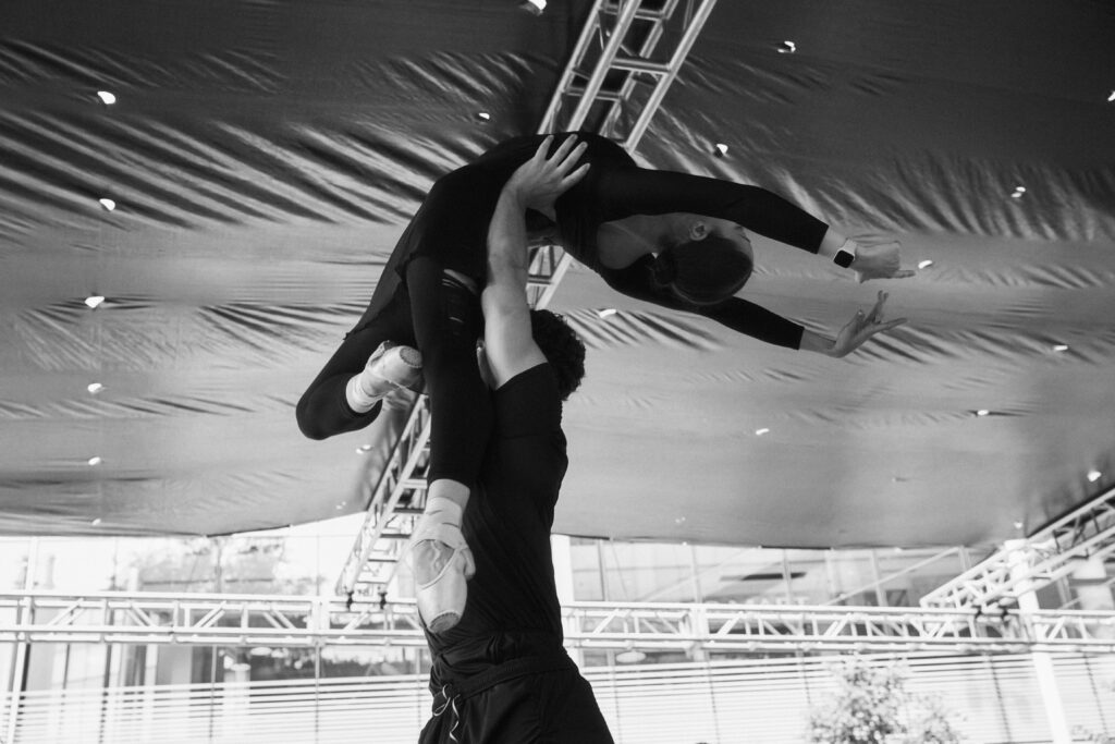 A black and white close-up of Logan Savard and Taisi Tollasepp partnering on an outdoor stage. Savard lifts Tollasepp in a press lift above his head. Tollasepp is arching backward, her face toward the ceiling and arms in fifth. One of her legs is extended straight down, the other in retiré. Both dancers wear all-black. Tollasepp wears a long sleeve leotard with a skirt, tights rolled at her ankles, and light pointe shoes. Savard wears a T-shirt and pants.