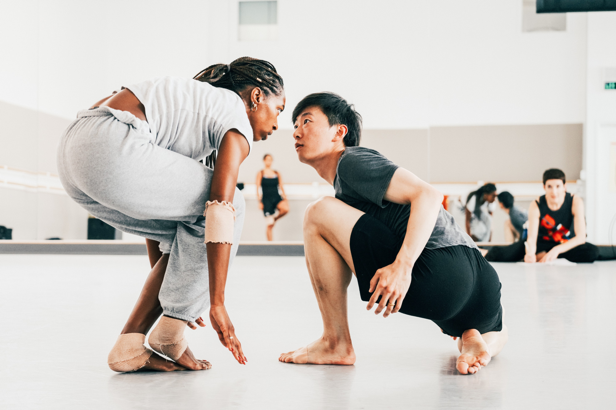 In a light-filled dance studio, Jacey Gaiilard, wearing gray sweatpants and a gray t-shirt, crocuhes down, her eyes fixed on Wei Wang. Wang, wearing a black t-shirt and shorts, squats low to the ground and looks up at her.