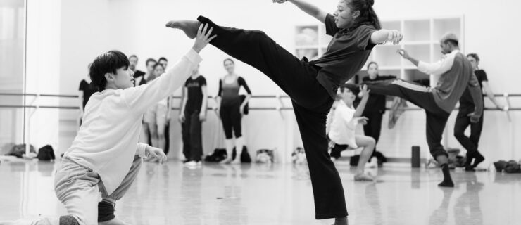 A black-and-white photo of dancers rehearsing a contemporary ballet in a large studio. Closest, one dancer kneels with one arm on his propped knee and his other reaching forward so his hand touches his partner's ankle. The second dancer lifts one leg front, slightly bent, with her arms to the side and her torso leaning back, concave in the upper chest. Other dancers rehearse or watch behind them.