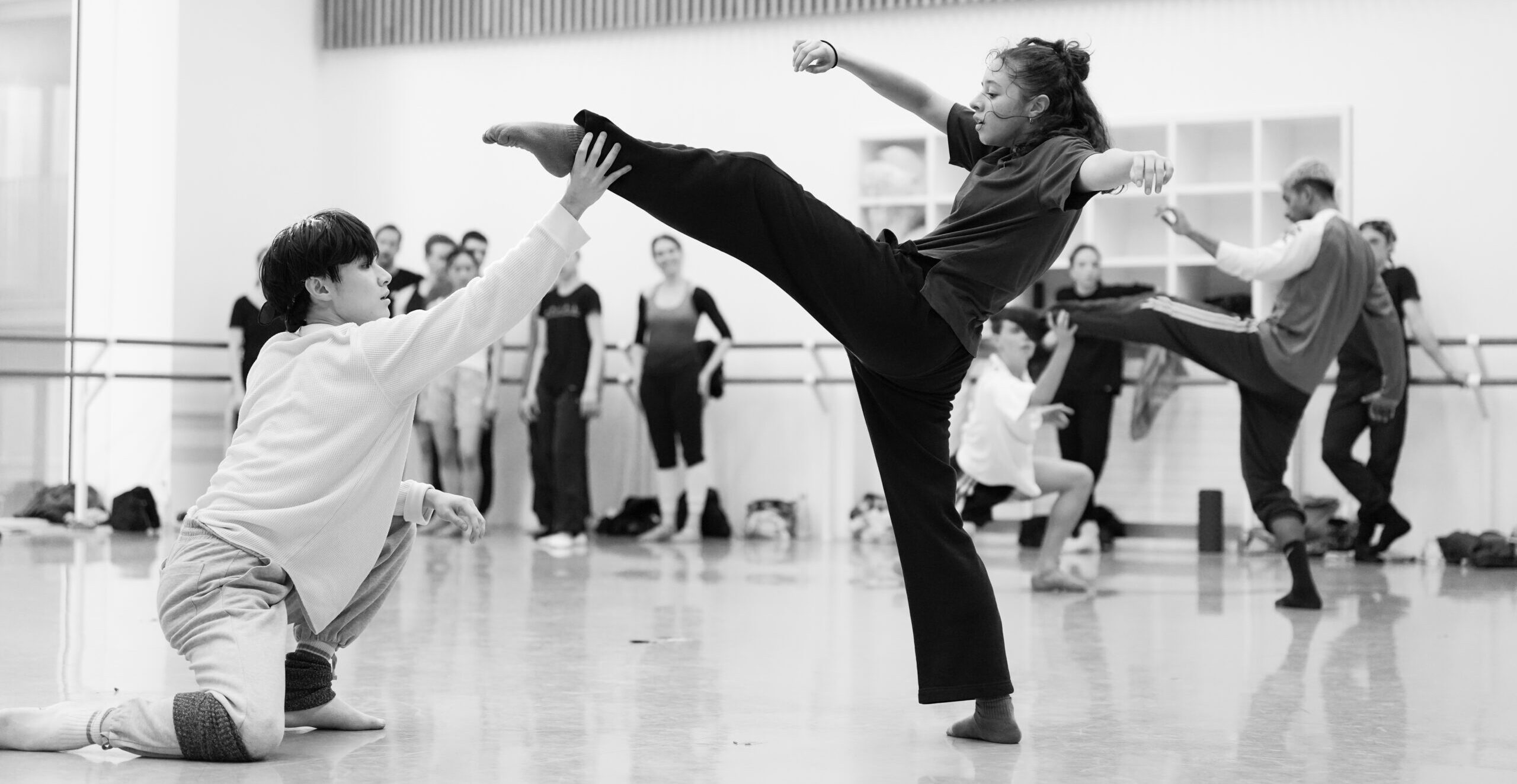 A black-and-white photo of dancers rehearsing a contemporary ballet in a large studio. Closest, one dancer kneels with one arm on his propped knee and his other reaching forward so his hand touches his partner's ankle. The second dancer lifts one leg front, slightly bent, with her arms to the side and her torso leaning back, concave in the upper chest. Other dancers rehearse or watch behind them.