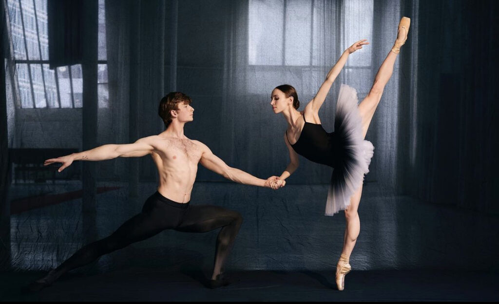 Aaron Bell and Skylar Brandt are shown dancing together in front of a dark grey translucent curtain in a shadowy dance studio. Bell, wearing black tights, is in a deep lunge facing Brandt with one arm extended behind him and the other out in front of him. Wearing a black ombré tutu and skin-tone pointe shoes, Brandt poses in penché en pointe, looking down at and holding Bell's outstretched hand.