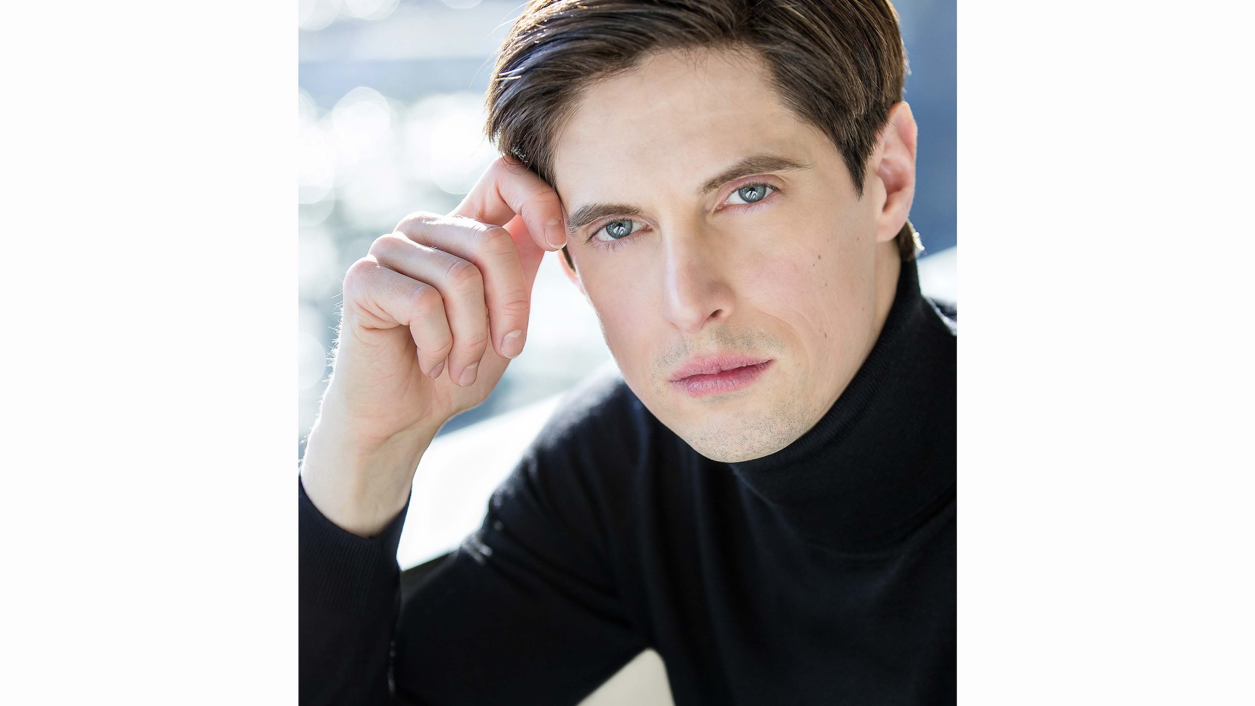 A headshot of Xander Parish, who wears a black turtleneck. He touches his temple with his right hand and looks directly towards the camera.