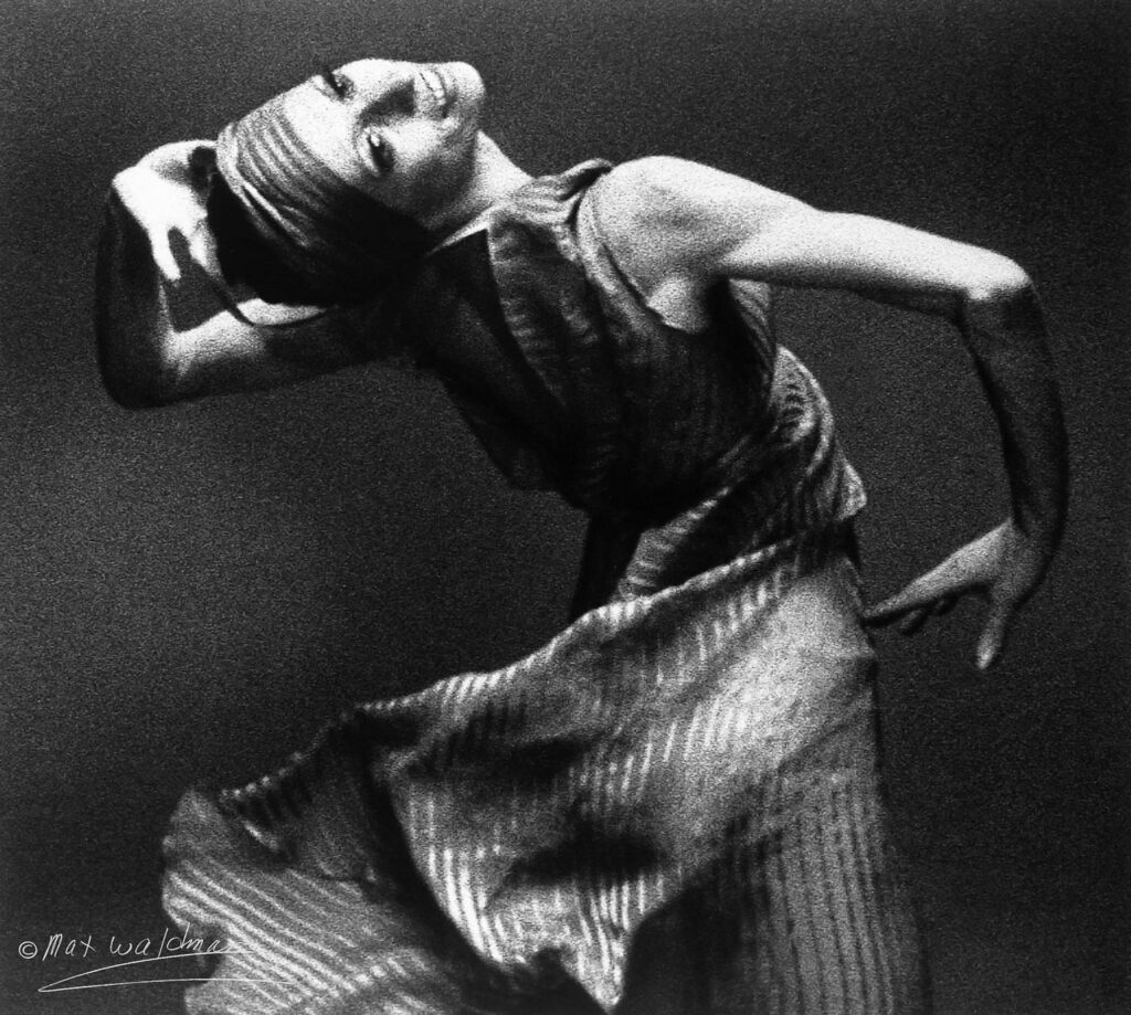 Natalia Makarova is shown from the thighs up as she twists her upper body to her left, with her left hand placed at the back of her head and her right hand on her right hip. She wears a striped, flowing dress and matching headscarf.