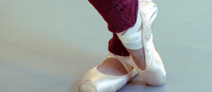 A close up photo of a dancer's lower legs and feet. She wears brugundy legwarmers and pointe shoes, and stands in parallel on her left foot as she crosses her right foot over her left ankle and presses over her toe.