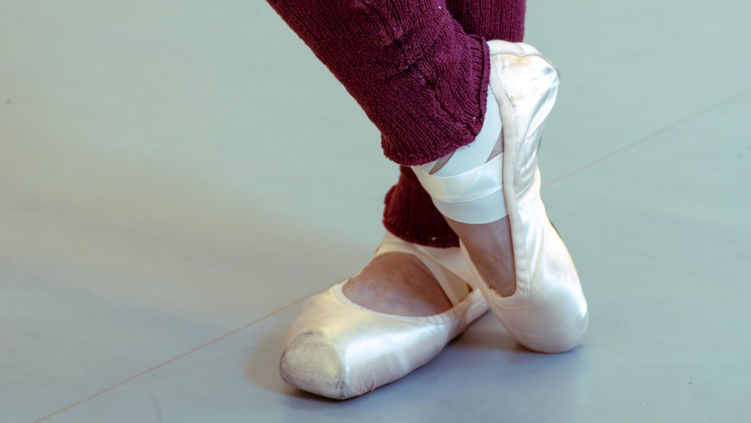 A close up photo of a dancer's lower legs and feet. She wears brugundy legwarmers and pointe shoes, and stands in parallel on her left foot as she crosses her right foot over her left ankle and presses over her toe.